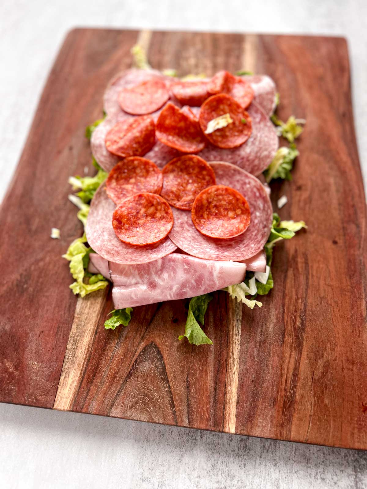 Salami, pepperoni, ham, and lettuce on a wood cutting board.